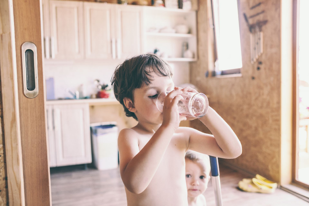 The boy drinks water from a glass. Children quench their thirst on a hot summer day. The brothers play together at home. Naked kids run around the house. The child is cleaning in the kitchen.